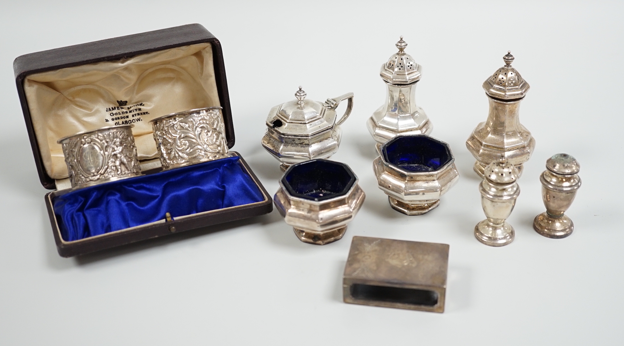 A cased pair of Edwardian embossed silver napkin rings, Birmingham, 1904, a five piece silver condiment set, Birmingham, 1926, two plated condiments and a match sleeve.